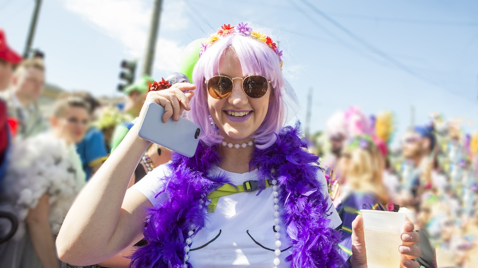 Costumed 20 something girl saying hello and holding phone surrounded by party goers